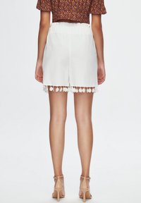 White shorts with a textured, elastic waistband and decorative lace trim. The hem features small fabric pom-poms. Flesh-toned heels are worn.