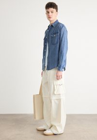 Džinsaina krekls ar pogu aizdari un divām krūšu kabatām, kopā ar krēmkrāsas cargo biksēm, kam ir sānu kabatas. Modelis tur brūnu rokassomu.