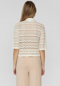 Lacejacka i krämfärgad, med zickzackmönster och korta ärmar, i en kort design. Mjuk textur och öppen väv.