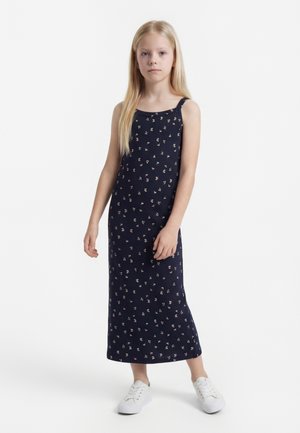 Mergina su ilgais šviesiais plaukais, vilkinti tamsiai mėlyną gėlėtą be rankovių suknelę ir baltus sportinius batelius, stovinti prieš baltą foną.