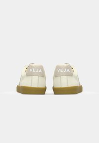 Hvite lær-sneakers med beige detaljer og gummisåle. Har 'VEJA'-logo på hælen og en jevn tekstur. Enkelt design.
