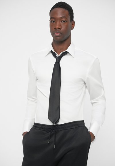 White long-sleeve shirt with a black tie; black drawstring pants. Smooth fabric, collared design, relaxed fit. Minimalist style.