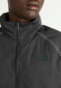 Gros plan d’une personne portant une veste noire zippée avec le logo « The North Face » sur la poitrine et une couture réfléchissante sur l’épaule.