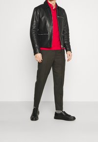Schwarze Lederjacke mit silbernen Reißverschlüssen, rotes Poloshirt, olivgrüne Anzughose und schwarze Sneakers mit grauen Akzenten und strukturierten Sohlen.