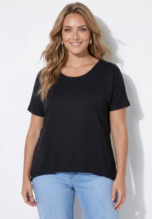 Lächelnde Frau mit welligem blondem Haar trägt ein schwarzes Kurzarmshirt und hellblaue Jeans, steht vor einem weißen Hintergrund.
