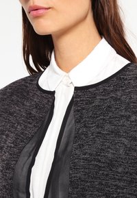 Svart och vit lagerblus med ett texturerat svart ytterlager och en genomskinlig tyg under, kompletterad med en vit knappkrage.