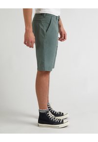 Pánské tyrkysové šortky vyrobené z bavlny, s plochým předním dílem, bočními kapsami a přiléhavým střihem. V kombinaci s černými vysokými teniskami.