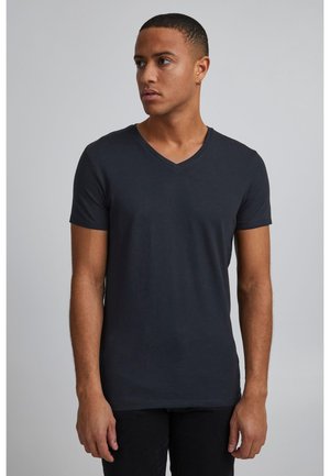 Νέος άνδρας που φοράει απλό μαύρο T-shirt με λαιμόκοψη σε σχήμα V, στέκεται μπροστά σε ανοιχτό γκρι φόντο, κοιτάζοντας ελαφρώς προς τα αριστερά.