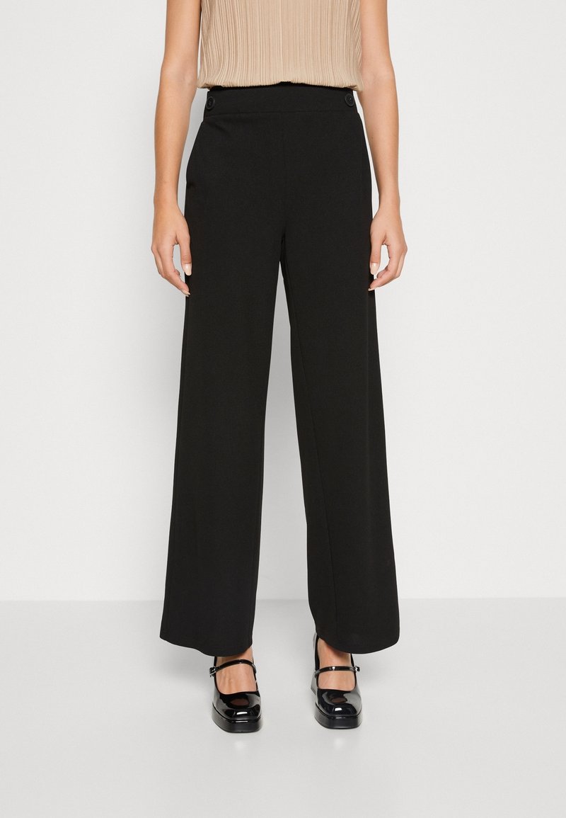 Femme portant un pantalon noir taille haute à jambes larges avec des boutons à la taille et des chaussures Mary Jane noires, top plissé sans manches beige.