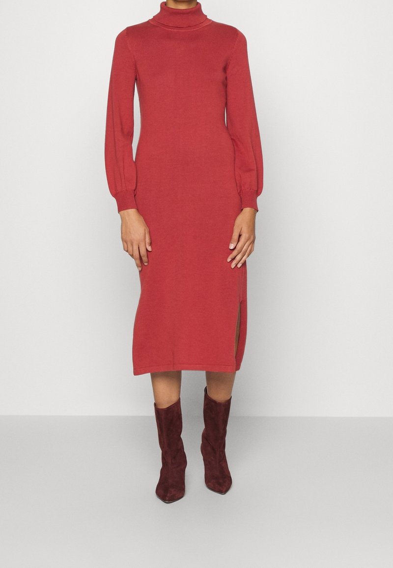 Personne portant une robe midi rouge à manches longues avec col roulé, une fente latérale et des bottines en suède rouge foncé, se tenant devant un fond uni.