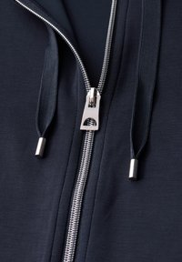 Námořnická bunda na zip se stříbrným metalickým zipem, texturovanými poutky na zatažení a hladkým materiálem, který zdůrazňuje jemné detaile stehu.