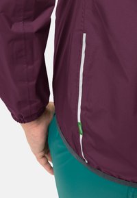Personne portant une veste violette avec une bande réfléchissante et un pantalon vert, la main reposant sur la hanche devant un fond blanc uni.