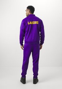 Fato de treino roxo com o texto amarelo "LAKERS" nas costas. Textura suave, corte atlético, inclui calças combinadas e ténis pretos.