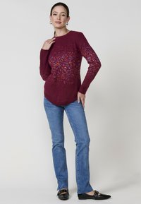Burgundy pulóver textúrált mintával, kerek nyakkal és hosszú ujjakkal, világoskék egyenes szárú farmernadrággal és fekete lapos cipővel párosítva.