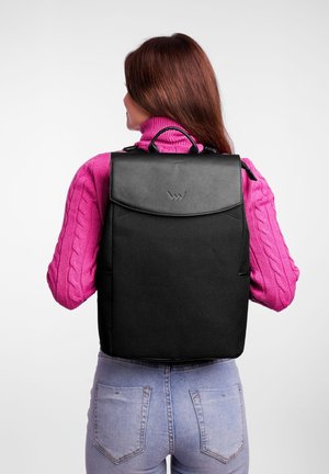 Schwarzer Rucksack mit einem Lederdeckel und Logo, der ein strukturiertes Design und verstellbare Träger aufweist, getragen mit einem rosa Pullover.