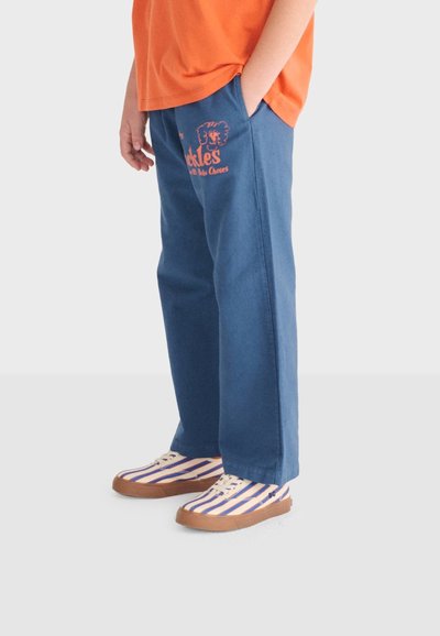 Enfant portant un pantalon bleu avec un texte orange et un motif d'éléphant, un t-shirt orange et des baskets blanches avec des rayures bleues et des semelles marron.