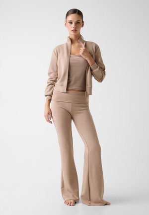 Femme en veste zippée beige, débardeur assorti et pantalon large, debout pieds nus devant un fond uni.