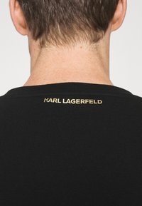 T-shirt nera in cotone con texture a coste e collo rotondo, dotata del marchio dorato "KARL LAGERFELD" sulla parte posteriore del collo.