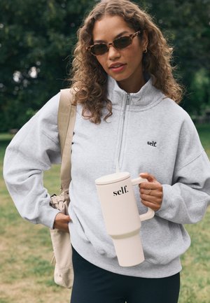 Naine lokkis juustega, kes kannab kilpkonnakujulisi päikeseprille ja halli ühe neljandiku lukuga dressipluusi, hoides käes suurt beežikat kruusi, millel on silt „self.”