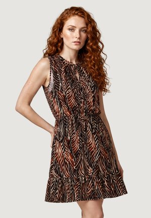Donna con lunghi capelli ricci rossi che indossa un vestito marrone e nero senza maniche con un orlo arricciato, in piedi con una mano sui fianchi.