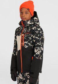 Schwarz-weiße gemusterte Schneejacke mit orangefarbenen Akzenten, ausgestattet mit einem hohen Kragen, Reißverschlusstaschen und Ärmelbündchen über den Handschuhen.