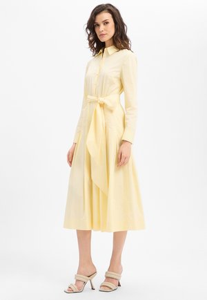 Frau trägt ein langärmeliges, blassgelbes Midikleid mit einer geschnürten Taubenschleife und beigen High-Heel-Sandalen, steht vor einem schlichten Hintergrund.