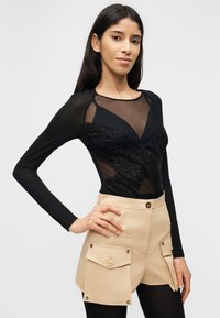 Γυναίκα με μακριά σκουρόχρωμα μαλλιά φορά διαφανές μαύρο τοπ με μαύρες λεπτομέρειες και μπεζ σορτς με μεγάλες μπροστινές τσέπες και κουμπιά.