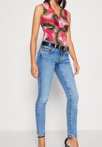 Woman wearing sleeveless red tropical print top, light blue skinny jeans with black belt, and black high heel shoes.