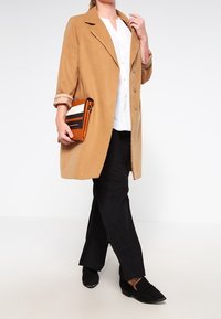 Beige kappa med notchade slag, knappstängning och upprullade ärmar, i kombination med svarta byxor och svarta slip-on skor. Orange randig clutch.