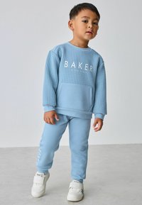 Lyseblå sweatshirt med struktureret front og hvid "BAKER" tryk, parret med matchende sweatpants og hvide sneakers.