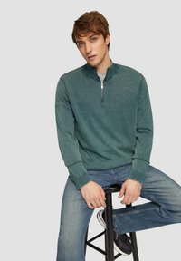 Donker teal pullover met een kwart ritssluiting, ribbelmanchetten en kraag. Draagt over een grijs shirt, gecombineerd met blauwe jeans en zwarte sneakers.