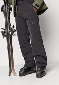 Sötétszürke sí nadrág, tartós anyagból, megerősített térdrésszel. Logó részlettel rendelkezik, és fekete sícipőkkel és sílécekkel van párosítva.