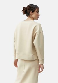 Beige cropped sweatshirt with a rounded neckline, dropped shoulders, and ribbed cuffs, featuring minimal branding on the back.
