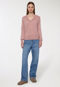 Jasnoróżowy sweter z dzianiny z dekoltem w serek i prążkowanym wykończeniem, w zestawie z luźnymi niebieskimi dżinsami i brązowymi butami wsuwanymi.