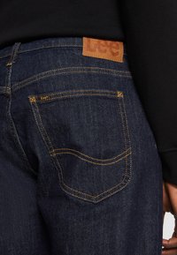 Vaqueros de mezclilla azul oscuro con costuras visibles y un parche de cuero en la parte trasera con el logo "Lee". El diseño del bolsillo incluye una forma curva y detalles dorados.