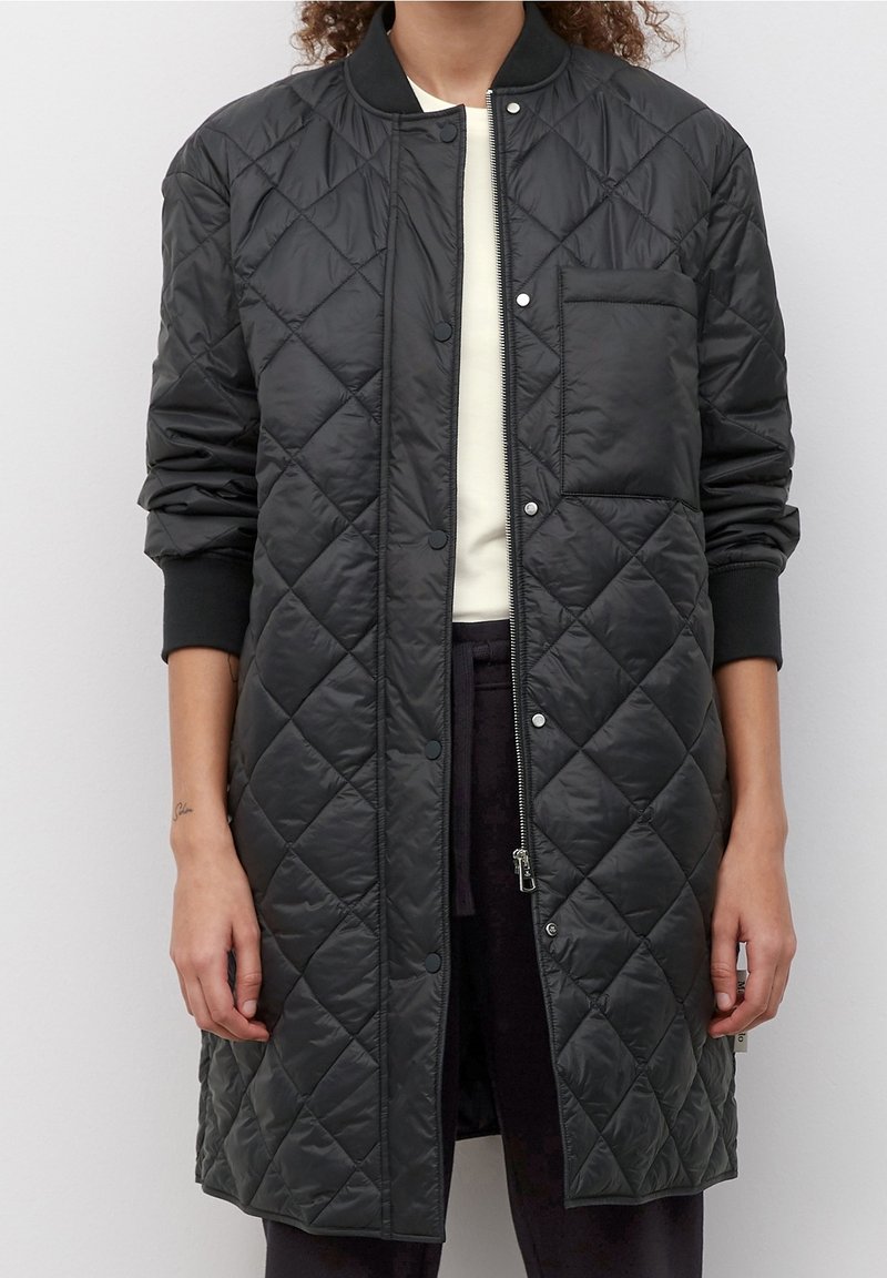 Chaqueta negra acolchada con cierre de cremallera y press-stud, puños acanalados y un bolsillo en el pecho. Presenta una textura suave y brillante.