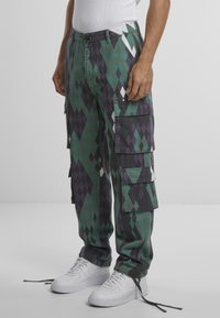 Pantalones cargo en un patrón de diamantes que presentan tonos verdes, negros y morados. Incluyen múltiples bolsillos y costuras reforzadas. Se llevan con zapatillas blancas.