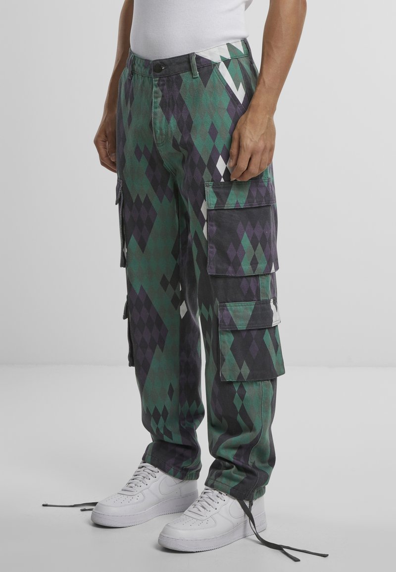 Pantalones cargo en un patrón de diamantes que presentan tonos verdes, negros y morados. Incluyen múltiples bolsillos y costuras reforzadas. Se llevan con zapatillas blancas.
