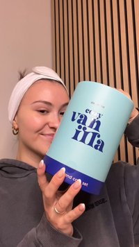 Een cilindrische, lichtblauwe container met het label "cozy vanilla", met donkerblauwe accenten en een matte afwerking, vastgehouden door een persoon in een grijze trui.