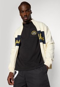 Kremfarget zip-up jakke med marineblå og gull detaljer, båret over en svart t-skjorte med et sirkulært merke. Modellen står i en nøytral posisjon.