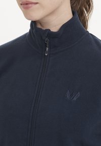 Veste en polaire navy avec un col haut et une fermeture éclair, présentant un logo brodé texturé sur la poitrine et une apparence douce et moelleuse.