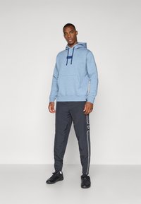 Lyseblå hættejakke med en frontlomme og marineblå snørebånd, parret med sorte bukser med hvide accenter. Sorte sko fuldender outfittet.