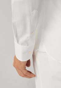 Dettaglio di una camicia bianca a maniche lunghe che mostra un polsino abbottonato, una texture liscia e una vestibilità sagomata, con un bottone visibile di un colore a contrasto.