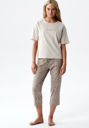 Młoda kobieta o długich blond włosach stoi boso, ubrana w beżową koszulkę z krótkim rękawem z napisem oraz jasnobrązowe, wzorzyste spodnie o długości 7/8.