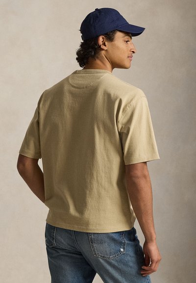 Μπεζ βαμβακερό t-shirt με κοντά μανίκια και άνετη εφαρμογή, συνδυασμένο με μπλε τζιν τζιν. Ένα σκούρο μπλε καπέλο ολοκληρώνει την εμφάνιση.