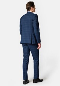 Granatowy garnitur w kratę, dopasowany krój. Posiada jeden rozcięcie i zapięcie na dwa guziki. Uzupełniony czarnymi butami.