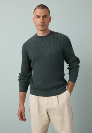 Suéter de canalé verde oscuro con cuello redondo y mangas largas, combinado con pantalones de color beige claro. Textura suave y ajuste relajado.