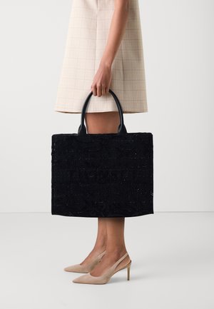 Grote zwarte tote bag met een textuur van een bloempatroon en logo detail, en stevige zwarte handvatten. Model staat met gekruiste benen en draagt beige hakken.