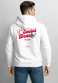 Weißer Hoodie mit "Social Beach Club" in roten und blauen Buchstaben auf der Rückseite, ausgestattet mit Sonnen- und Wellenmotiven. Die Vorderseite ist schlicht, der Stoff wirkt weich.