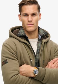 Sudadera con capucha de tela verde oliva con un cuello de forro polar, superpuesta sobre una camisa de franela a cuadros; presenta un reloj dorado en la muñeca.